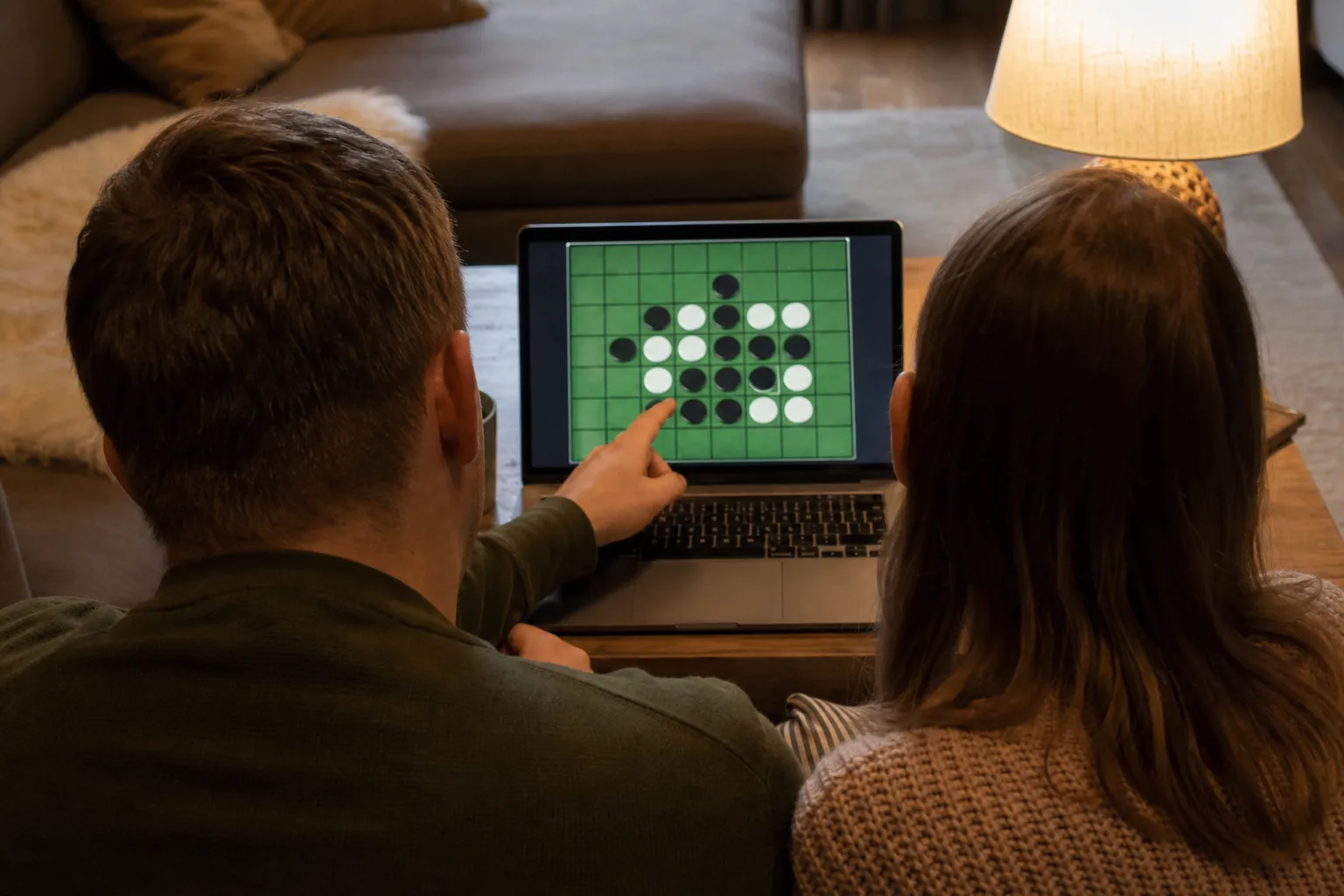 Two people sitting side by side, one pointing at a Reversi board on a laptop